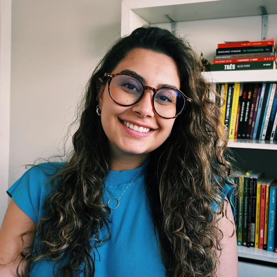 Retrato de Loreta Andrade sorrindo, de óculos de armação tartaruga, cabelos longos cacheados e blusa azul, diante de uma estante com livros.